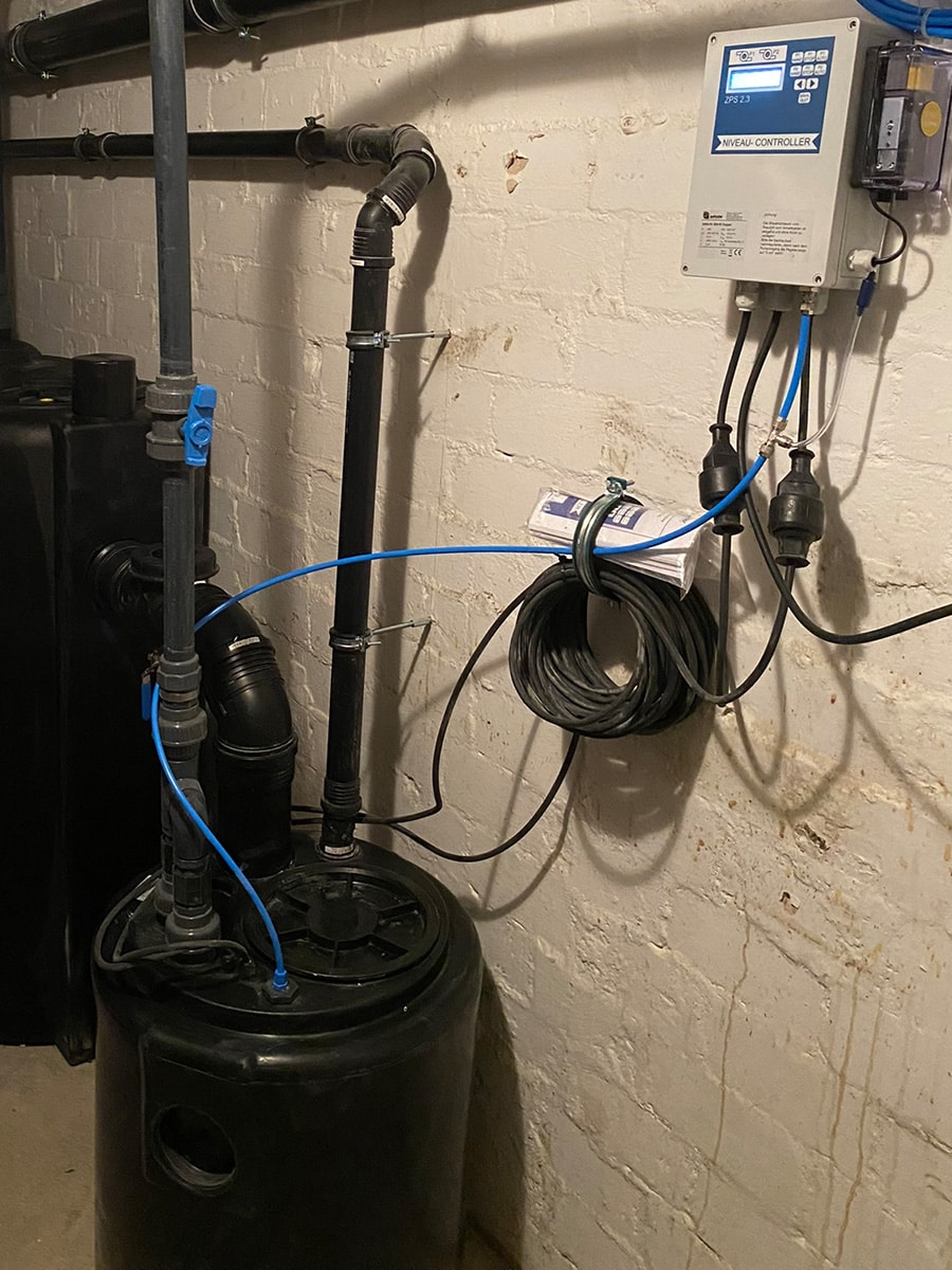 Fettabscheider und angeschlossene Hebeanlage im Keller mit schwarzen Rohrleitungen, blauen Leitungen und einem Steuergerät an der Wand